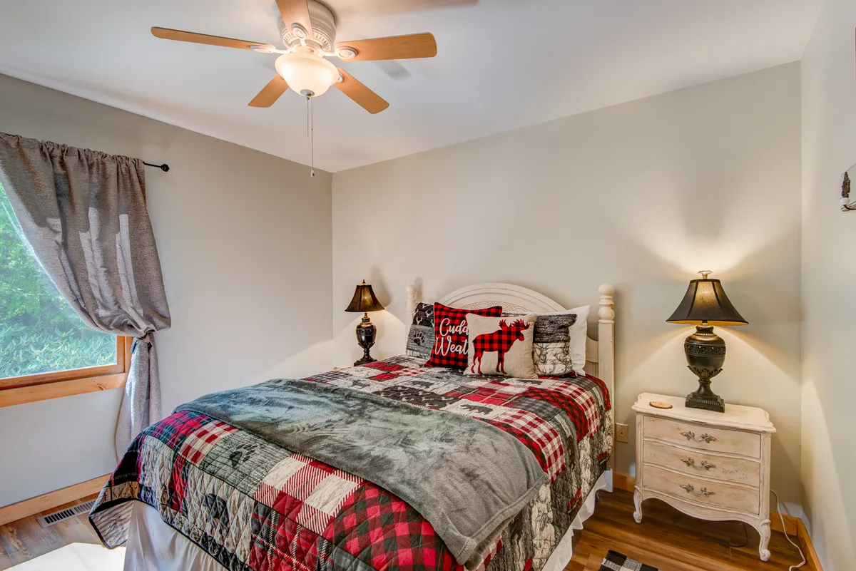 A restful bedroom with a patterned quilt, nightstand with lamp, and window framed by soft curtains. The decor is warm and inviting, with sunlight gently illuminating the space, creating a perfect environment for peaceful sleep or quiet moments.