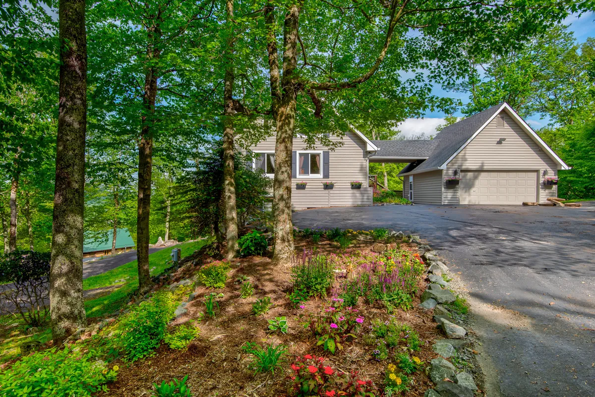 The image showcases the charming facade of a single-family mountain home surrounded by colorful flower beds, a spacious driveway, and mature trees. The tranquil setting is bathed in the soft light of sunrise, highlighting the inviting porch and lush, well-kept gardens, with hints of the Blue Ridge Mountains in the background.