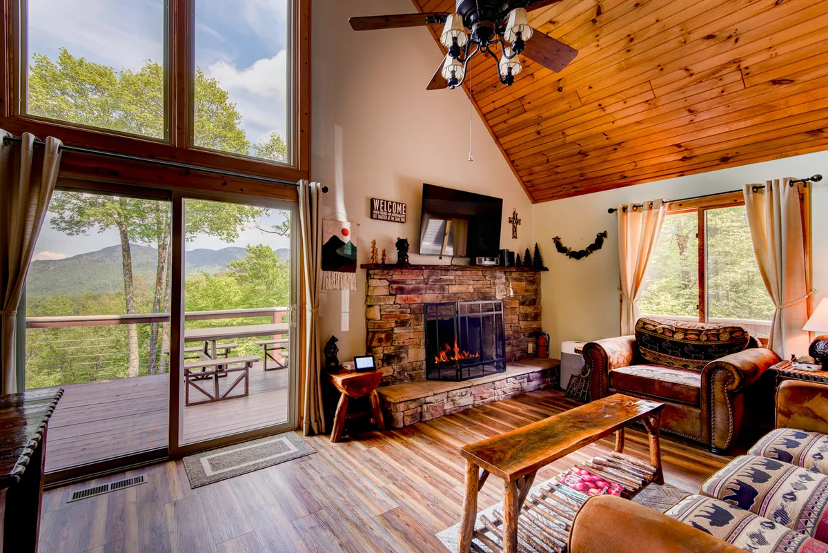 This interior image highlights a cozy seating arrangement around a prominent fireplace, complemented by large windows that reveal mountain scenery. Warm wood accents and decorative elements enhance the inviting, communal atmosphere of the living area.