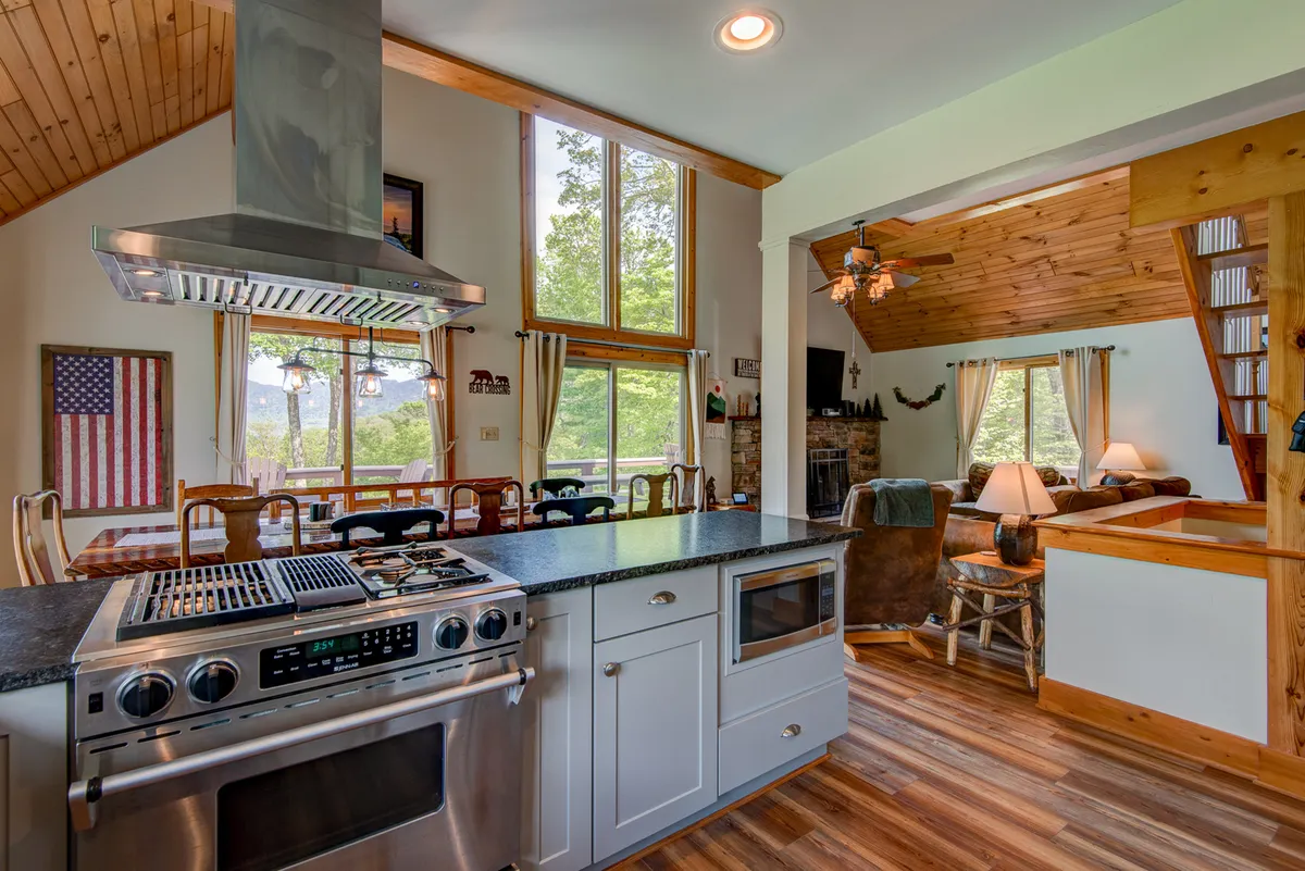 This image presents an open-plan interior scene featuring a kitchen island with stovetop, a large wooden dining table, comfortable seating, and expansive windows showcasing scenic mountain views. The space is filled with natural light, and the layout encourages both cooking and conversation, emphasizing the home's role as a place for gathering and entertaining.