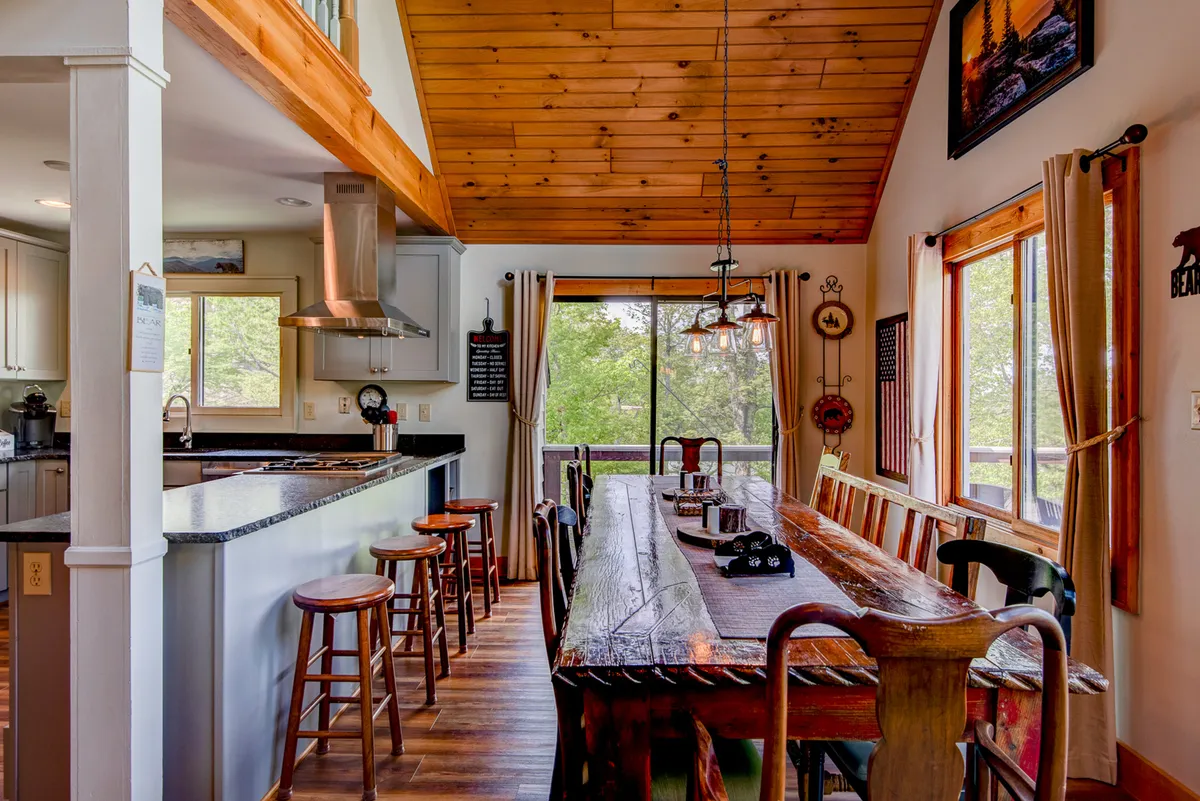 A spacious dining area is shown, featuring a large wooden table, exposed beams, a kitchen island, and expansive windows looking out to the mountains. The room is bathed in natural light, and the arrangement is perfect for family meals or celebrations with friends.