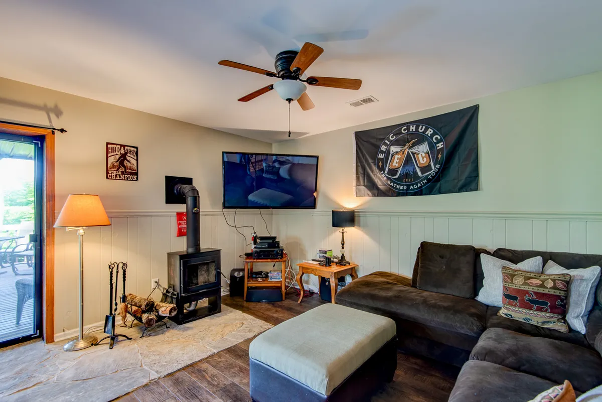 This inviting space features a sofa, a television, a wood stove, a ceiling fan, and a decorative flag on the wall. The setup encourages cozy gatherings for watching movies, socializing, or enjoying the warmth of the wood stove.