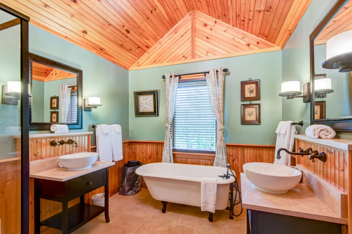 A spa-like bathroom featuring a freestanding soaking bathtub, dual vanities with sinks, a large window letting in abundant natural light, and a rustic wooden ceiling. The tranquil ambiance and high-end finishes evoke luxury and relaxation.