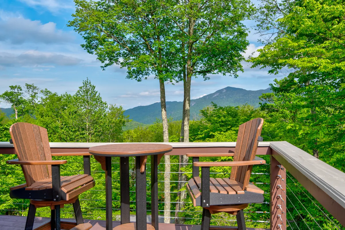 Wooden chairs and a small round table are positioned to face a breathtaking mountain view, with lush greenery enhancing the tranquil setting. The scene exudes peace and invites group members to share moments together, whether starting the day with coffee or unwinding in the evening.