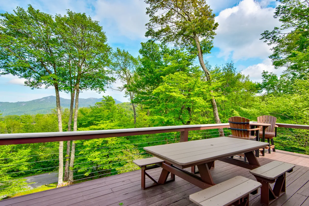 A wooden lower deck is featured with a picnic table and chairs, surrounded by tall trees and overlooking a scenic mountain view. The setting is peaceful, offering a perfect spot for group dining or quiet contemplation amid nature.