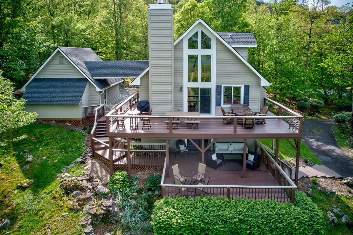 A spacious, elevated deck wraps around the house, featuring tall windows that frame scenic mountain views and let in abundant natural light. Outdoor seating and a stone pathway invite guests to gather, dine, or simply relax while surrounded by greenery. The deck is decorated for the season, suggesting a welcoming spot for both morning coffee and evening get-togethers.
