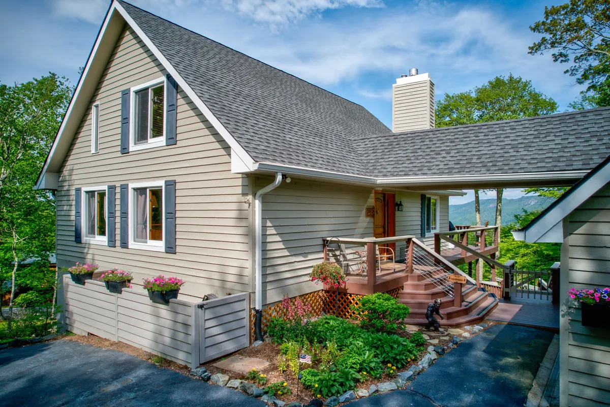 This image presents the welcoming exterior of the mountain home, featuring a charming front porch with flower boxes, a landscaped garden, and a visible chimney, all set against a tranquil backdrop. The inviting porch hints at the peaceful, private retreat described in the section.