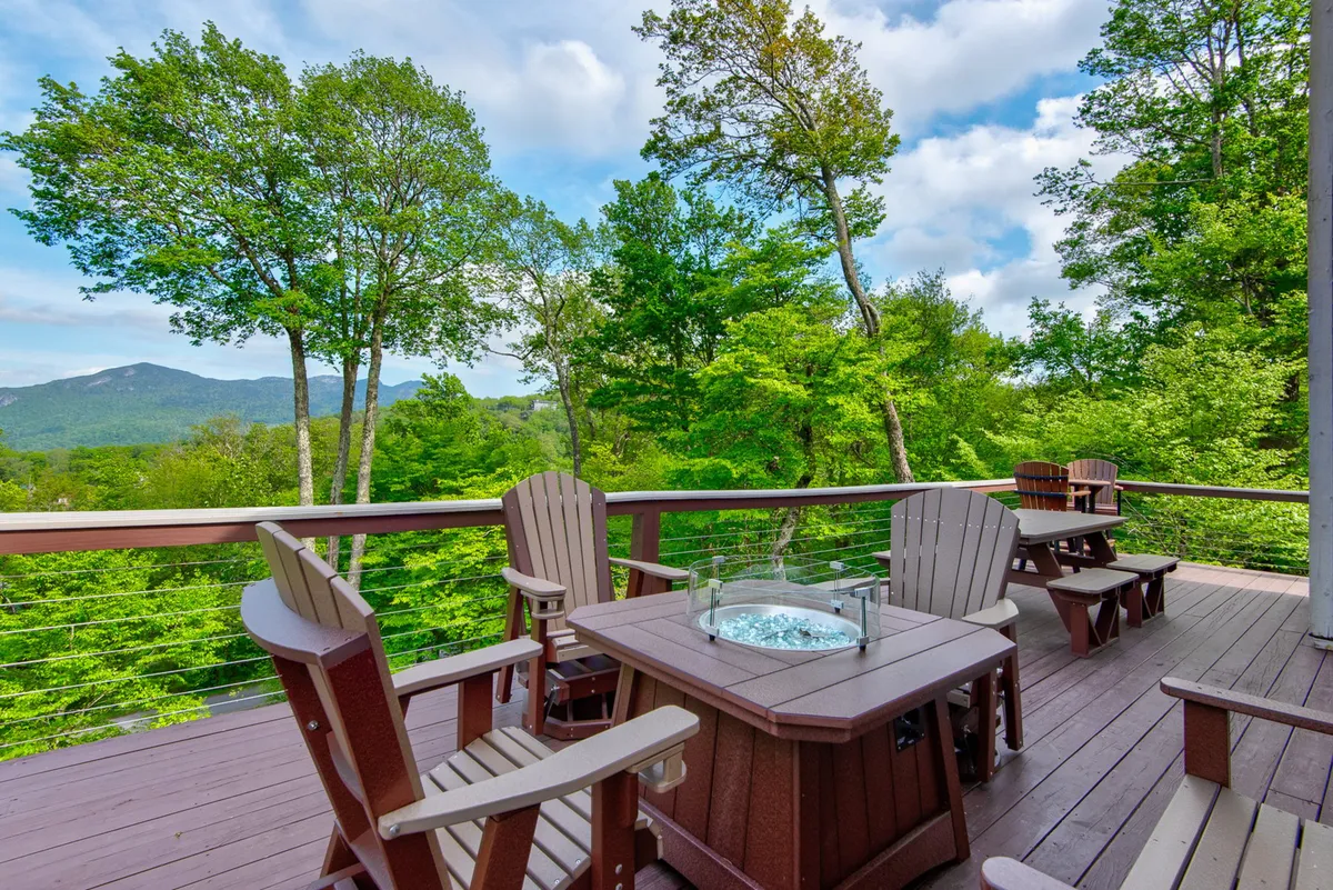 A spacious wooden deck is shown with comfortable seating arranged around a fire pit, all overlooking sweeping mountain views and lush greenery. The inviting setting suggests relaxation and social gatherings, with ample space and a tranquil atmosphere ideal for outdoor living.